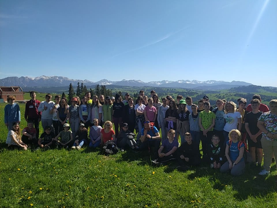 Uczniowie wraz z nauczycielami na zdjęciu grupowym na wzniesieniu Bachledówka w Czerwiennem koło Zakopanego. W tle panorama Tatr i Beskidu Żywieckiego. 