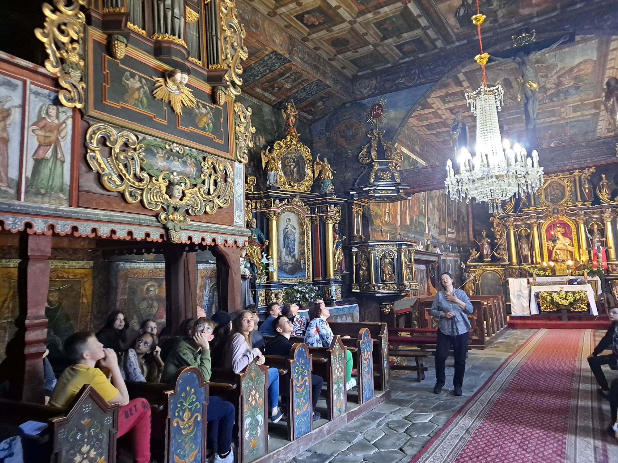 Uczniowie zwiedzają wnętrze zabytkowego kościoła św. Jana Chrzciciela w Orawce, siedzą w ławkach i podziwiają bogate malowidła świątyni, o których opowiada przewodnicza.