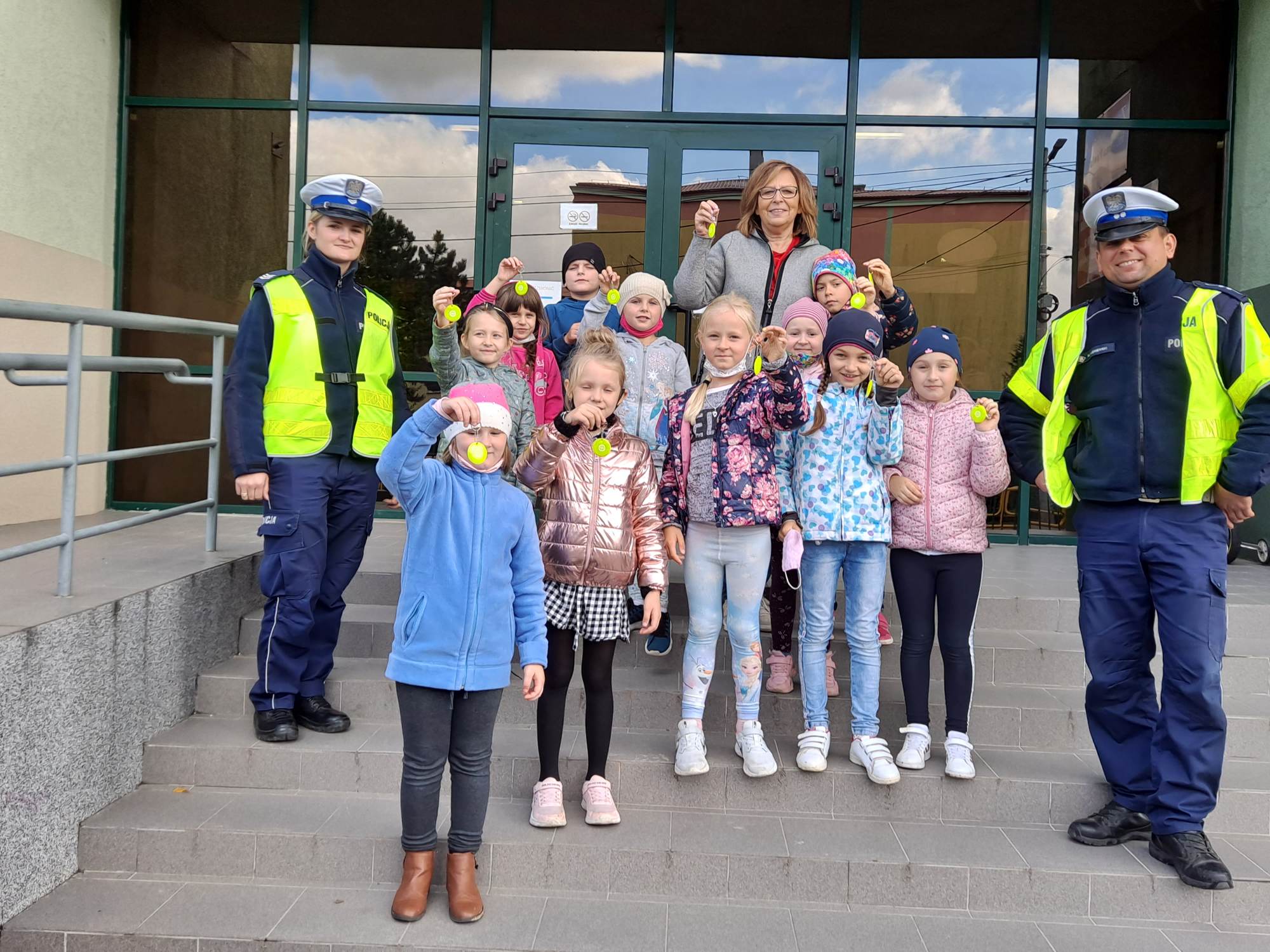 Dzieci trzymają breloczki - odblaski, stoją na schodach przed szkołą w towarzystwie policjantów ruchu drogowego