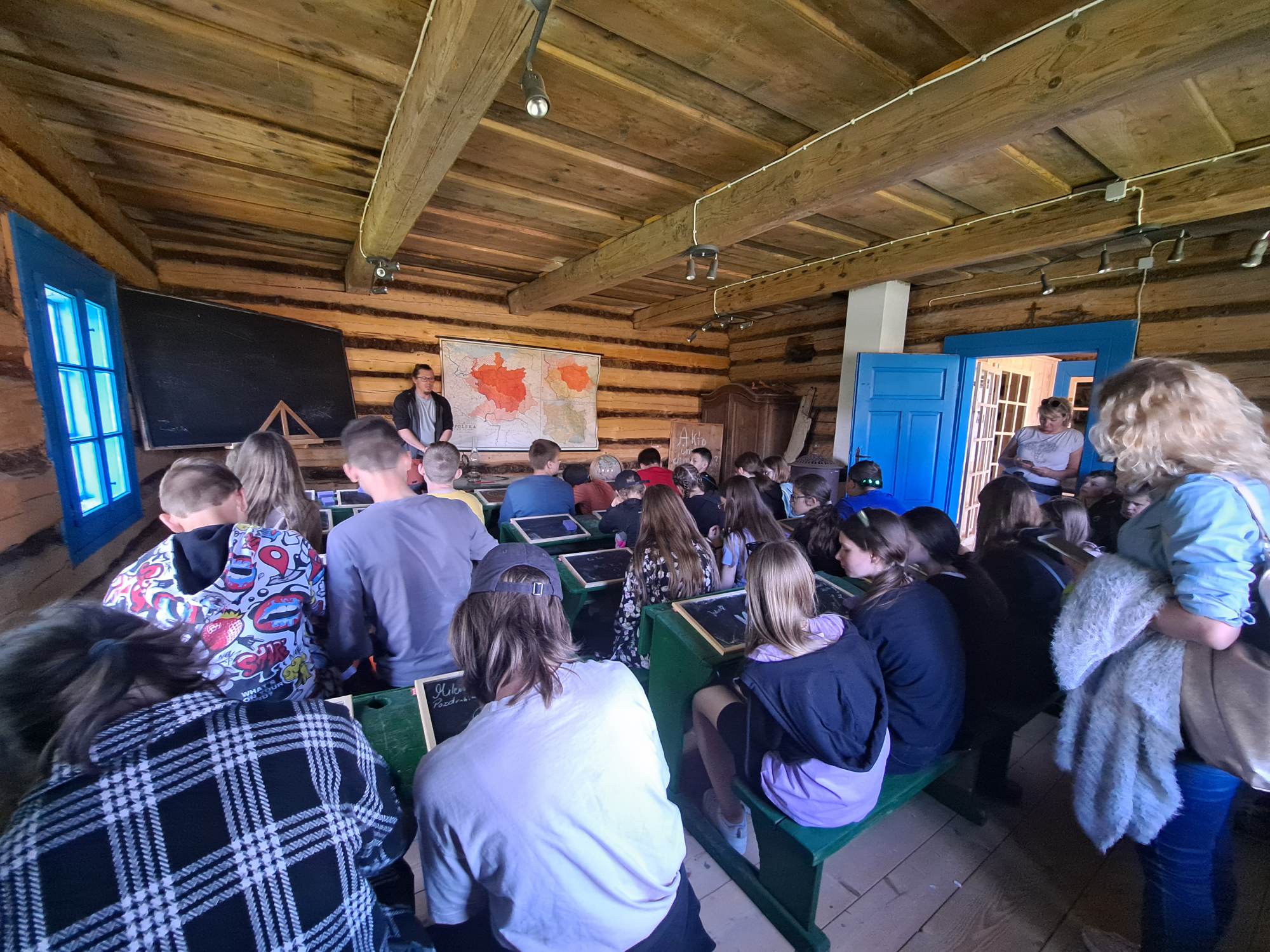 Uczniowie siedzą w ławkach dawnej szkoły znajdującej się w Muzeum - Orawskim Parku Etnograficznym, słuchają przewodnika i piszą kredą na tabliczkach.