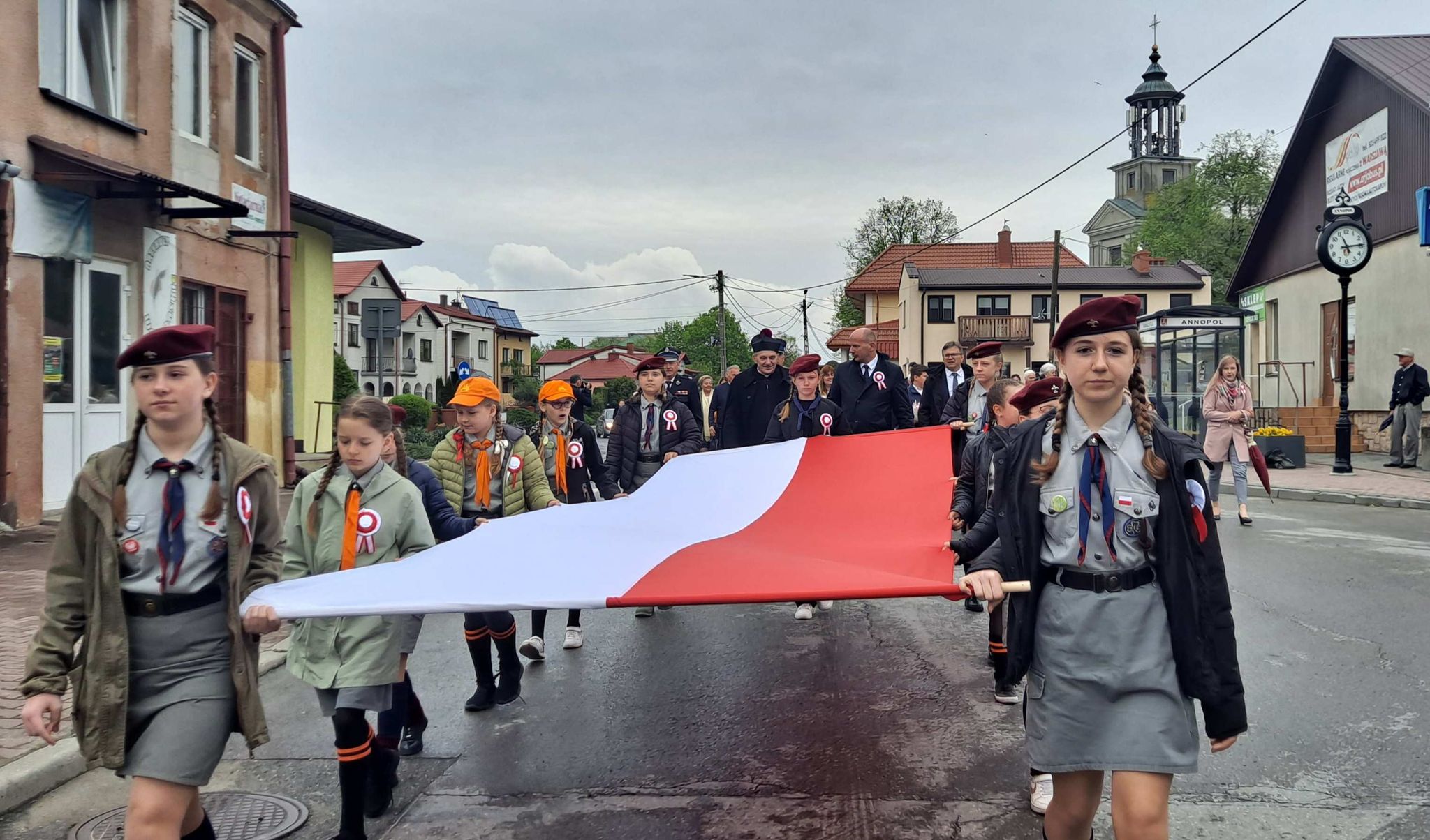 Zuchy i harcerze niosą biało-czerwoną flagę podczas gminnych uroczystości Święta Narodowego Trzeciego Maja