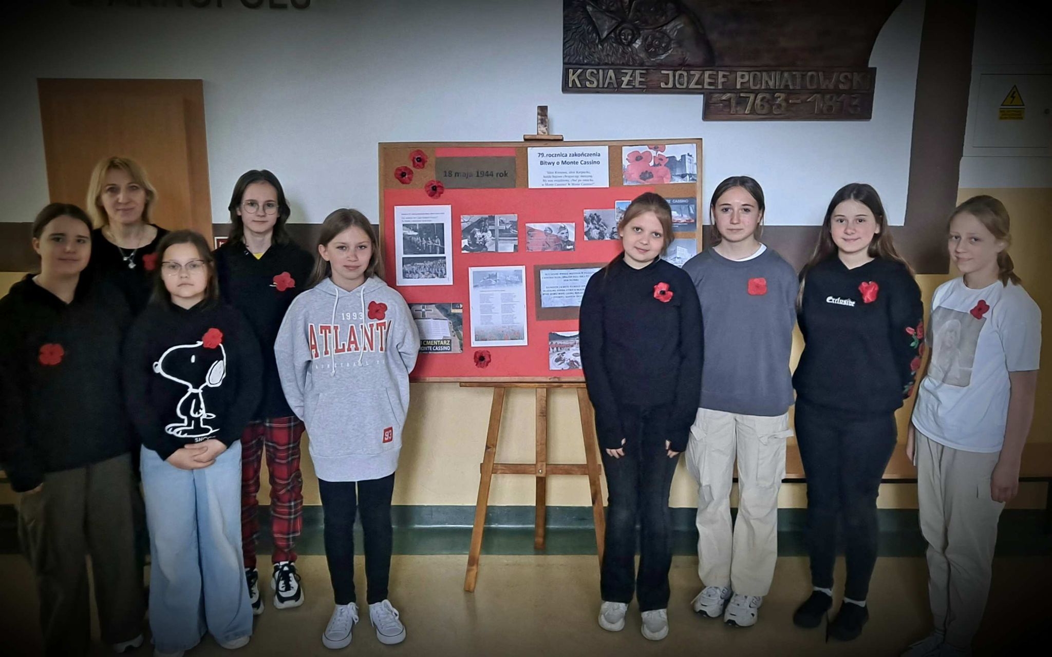 Dziewczęta z przypinkami na ubraniu - czerwonymi makami, przy gazetce tematycznej upamiętniającej rocznicę Bitwy o Monte Cassino, ustawionej na szkolnym korytarzu