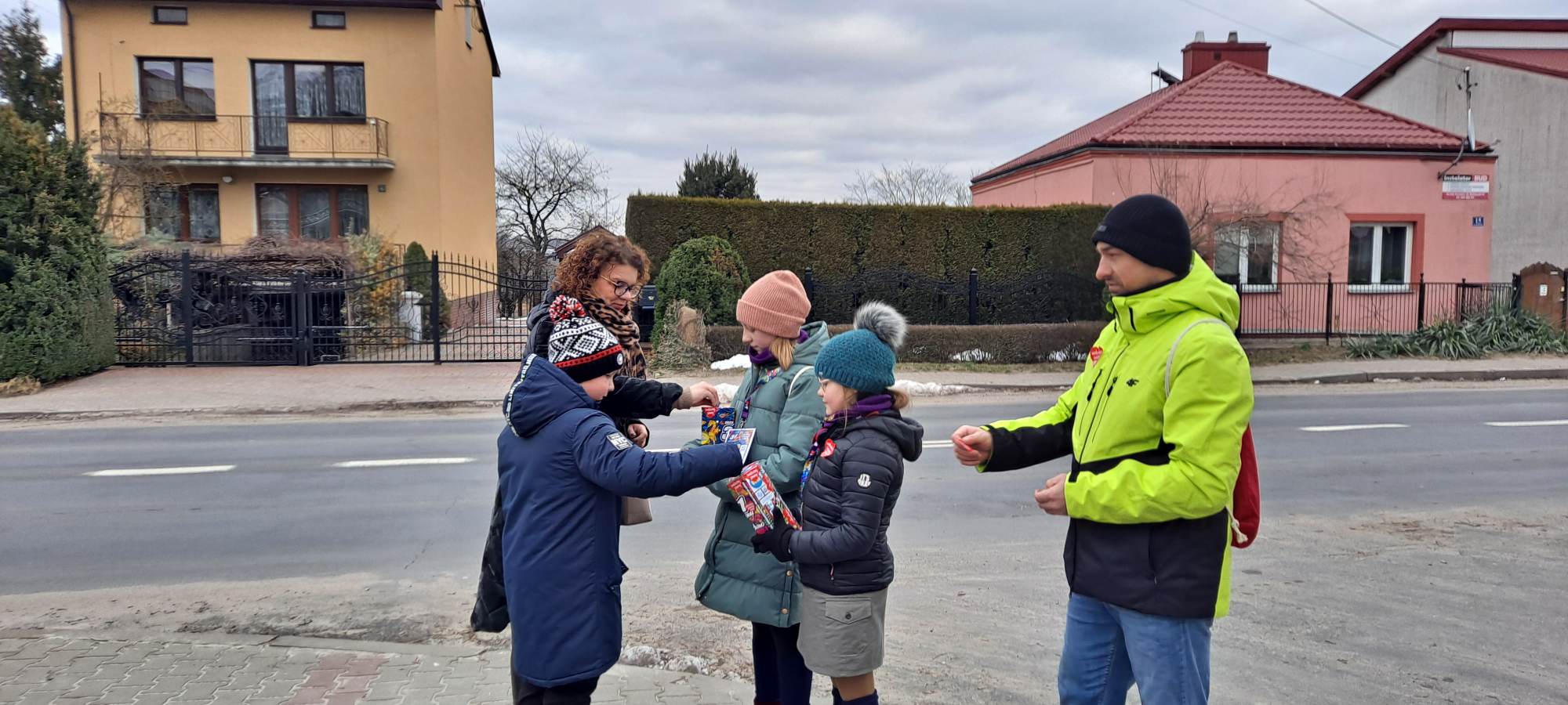 Harcerki - wolontariuszki 31.finału WOŚP wraz z opiekunem kwestują na ulicy miasteczka.Rodzic i dziecko wrzucają pieniądze do puszki.