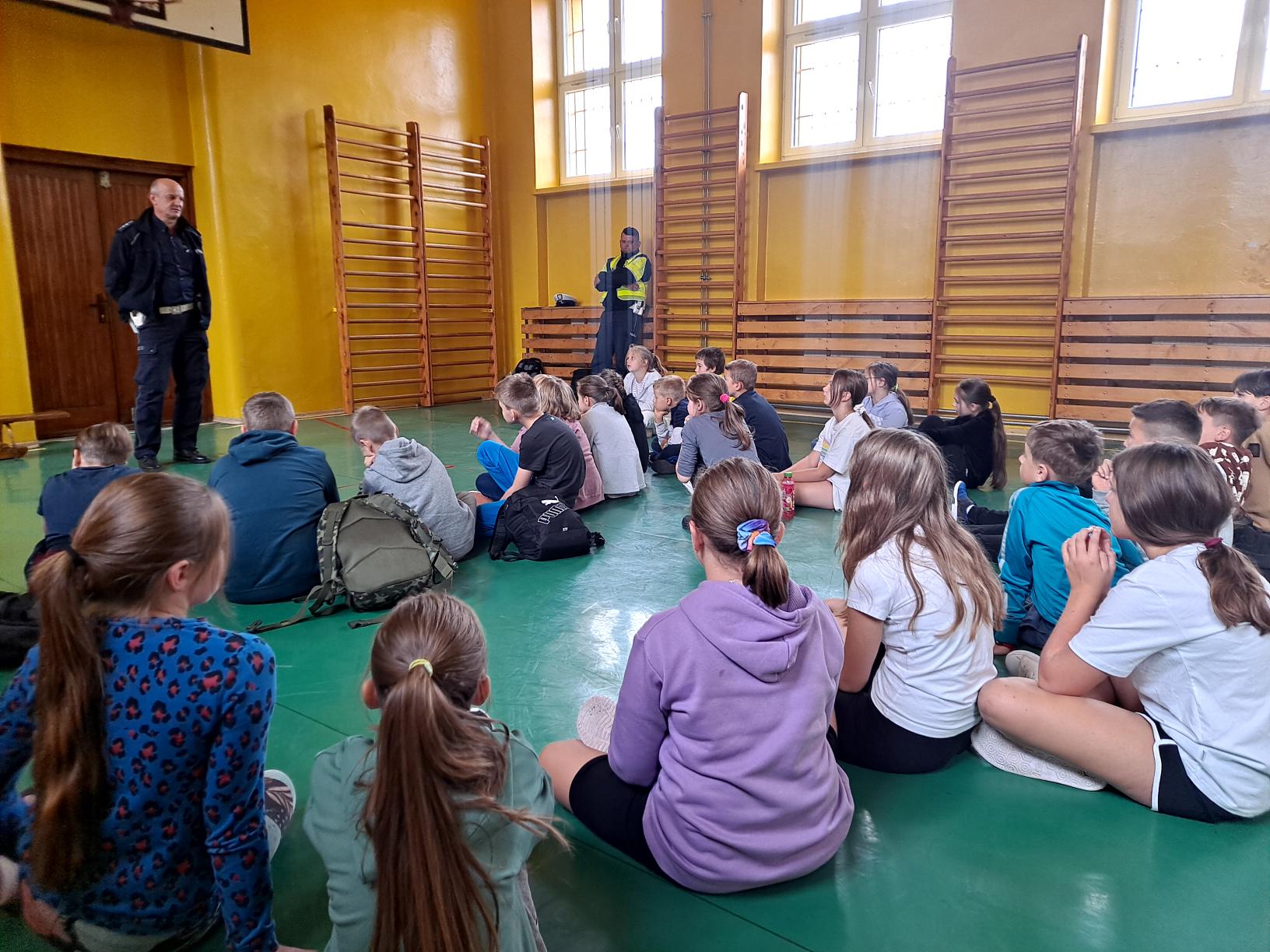 Uczniowie klasy czwartej uczestniczą w pogadance z policjantami z Wydziału ruchu Drogowego. Dzieci siedzą na podłodze w sali gimnastycznej, przed nimi dwóch policjantów. .